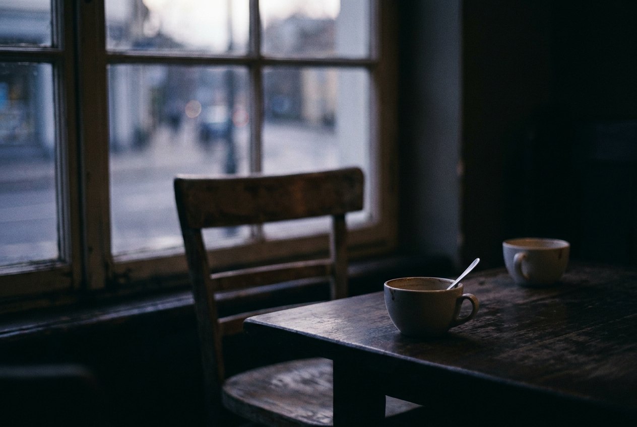 Two coffee cups on a dark table, one pushed away across an empty chair.